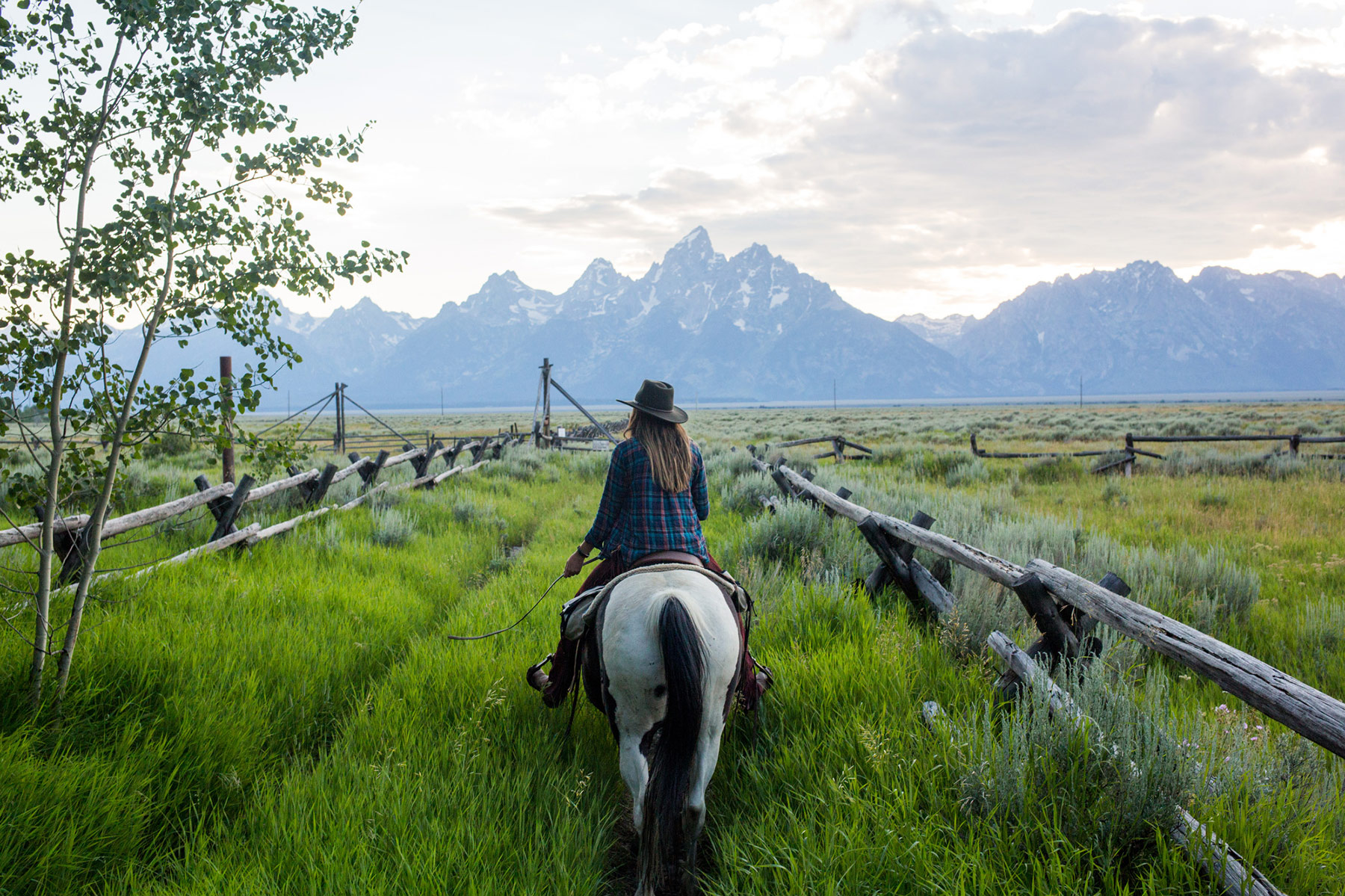A person wearing a hat and plaid shirt rides a horse along a grassy path bordered by rustic wooden fences in Idaho, with tall mountains visible in the distance under a partly cloudy sky. Photo by Sofia Jaramillo captures this unrivaled scene.