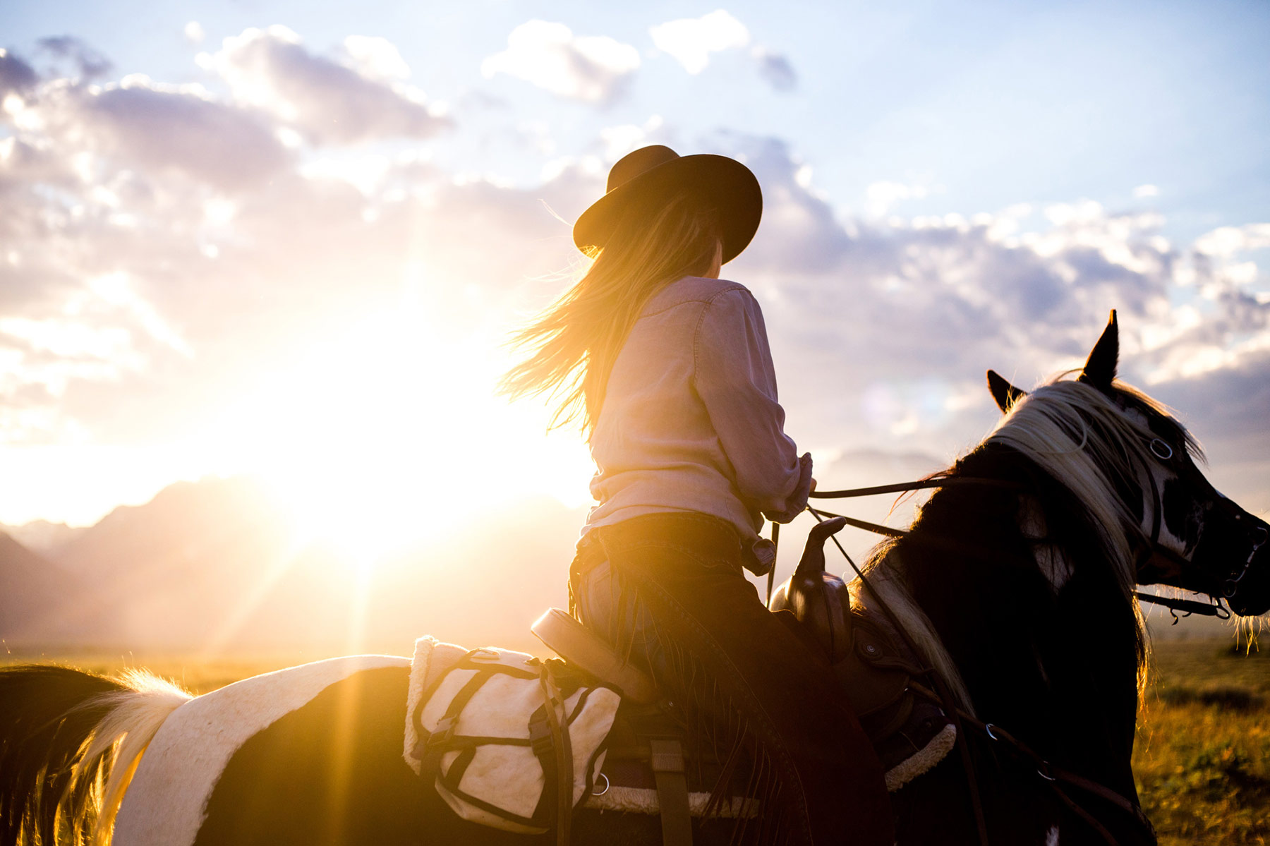 A person wearing a hat rides a horse in a field at sunset, with sunlight streaming in the background and clouds in the sky, capturing the essence of an outdoor adventure photographer’s perfect moment.