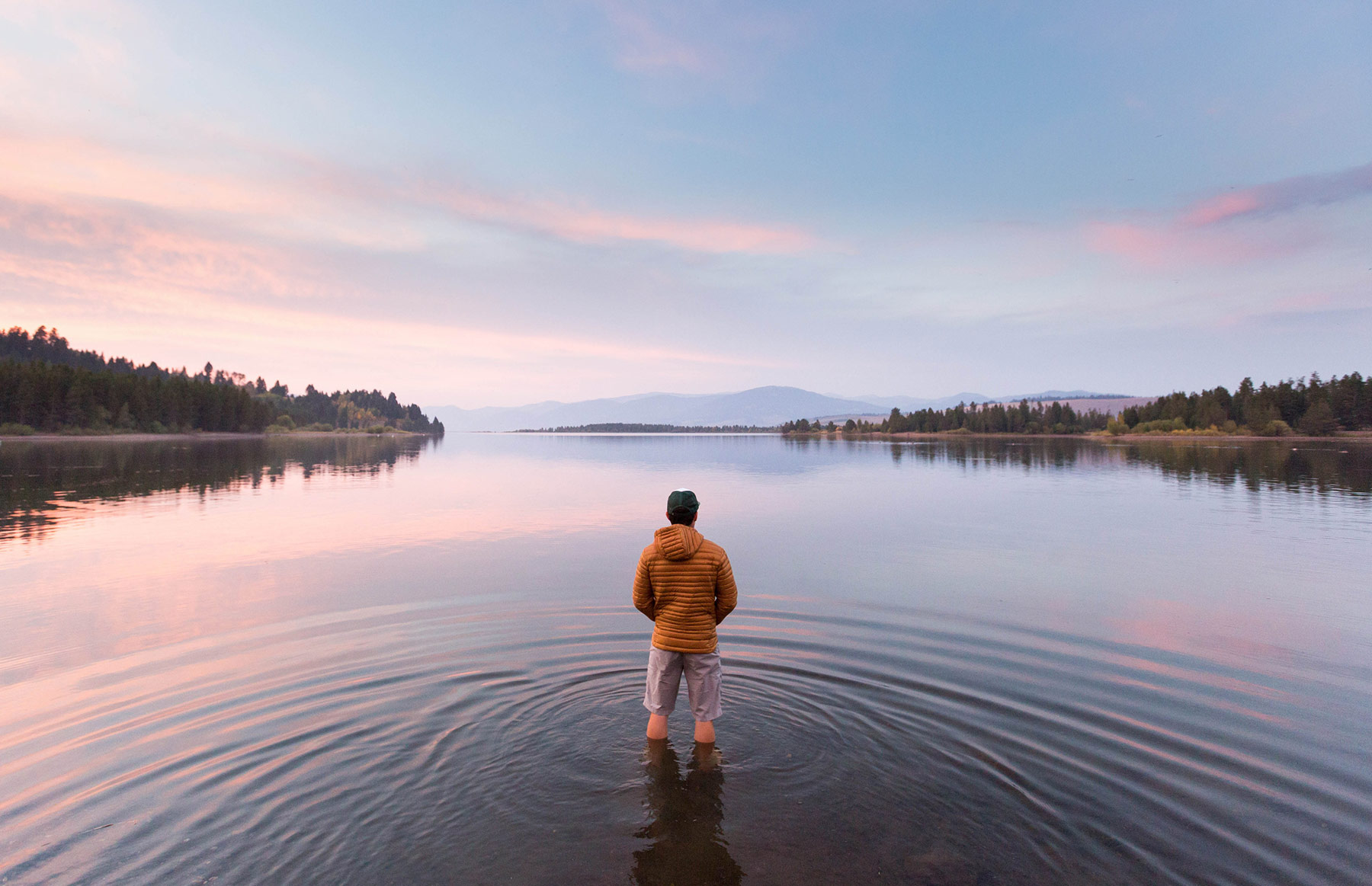 A person in a yellow jacket, clad in outdoor apparel, stands in shallow water at the edge of a calm lake, surrounded by trees and mountains in Idaho, under a pastel-colored sky at sunset or sunrise.