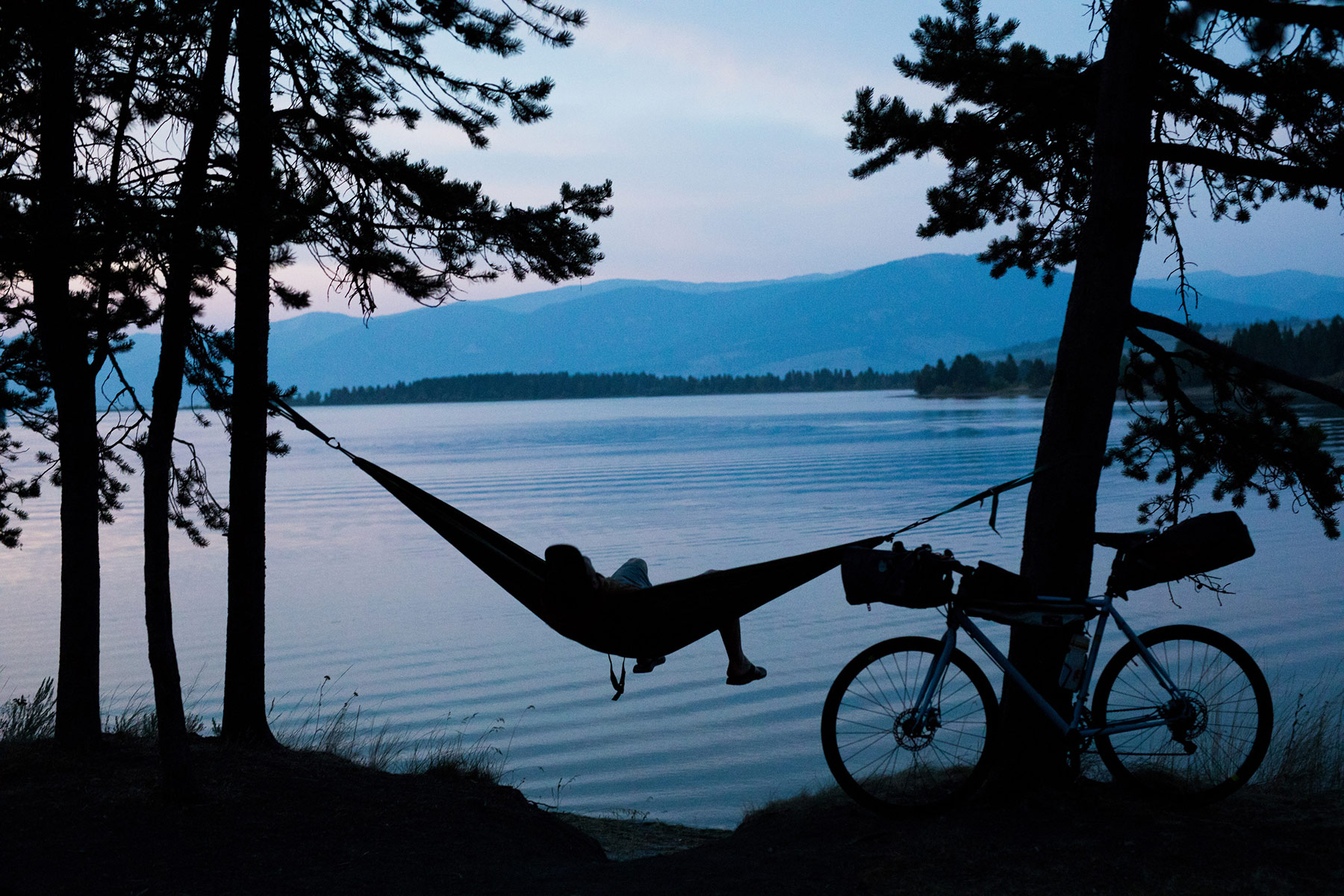 A person relaxes in a hammock tied between two trees by a lakeshore at dusk in Idaho, with a bicycle parked nearby and mountains visible—capturing the spirit of outdoor adventure photographer Sofia Jaramillo.