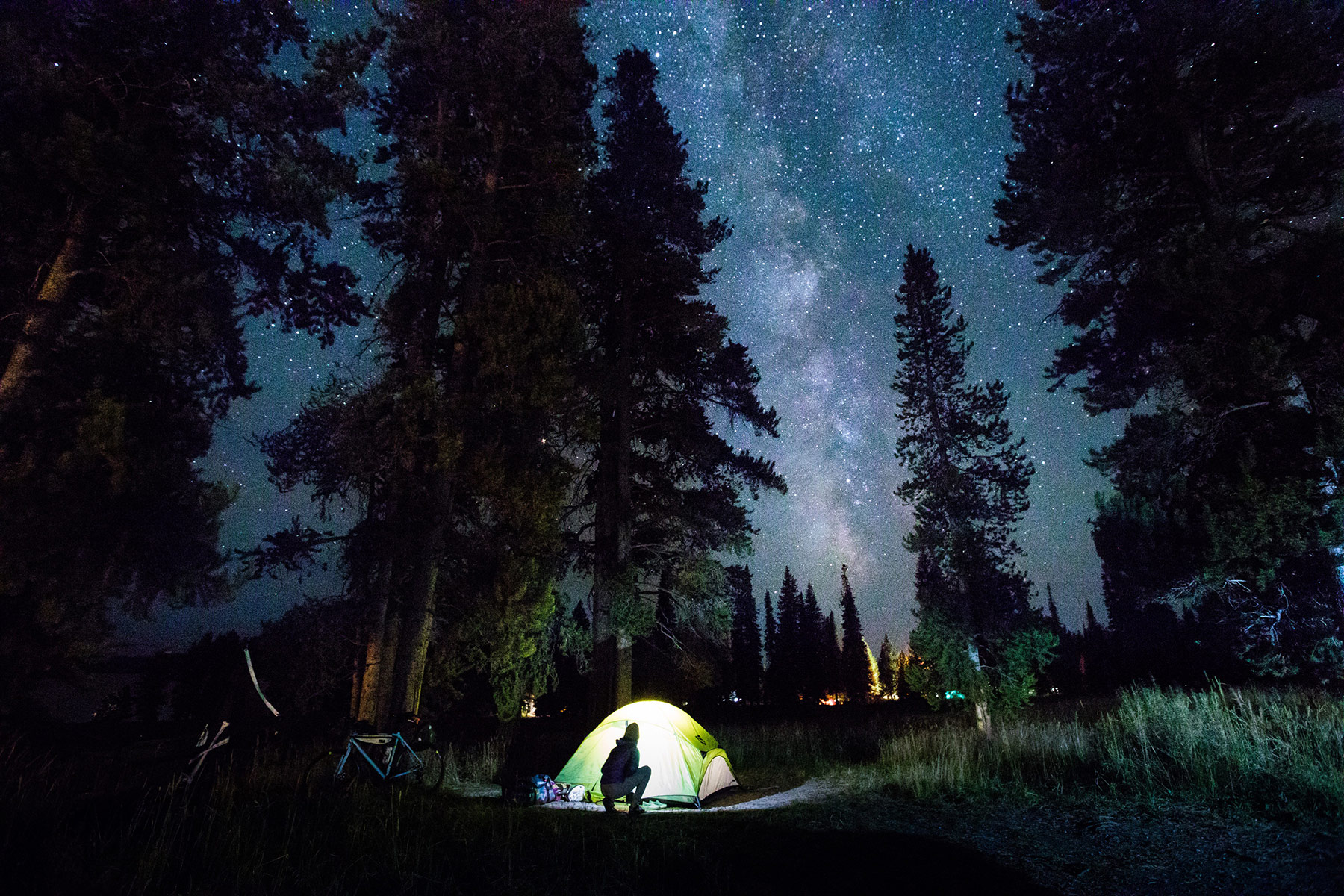 A person sits outside a glowing tent, surrounded by tall trees, under a clear night sky filled with stars and the Milky Way, enjoying the outdoors in cozy outdoor apparel.