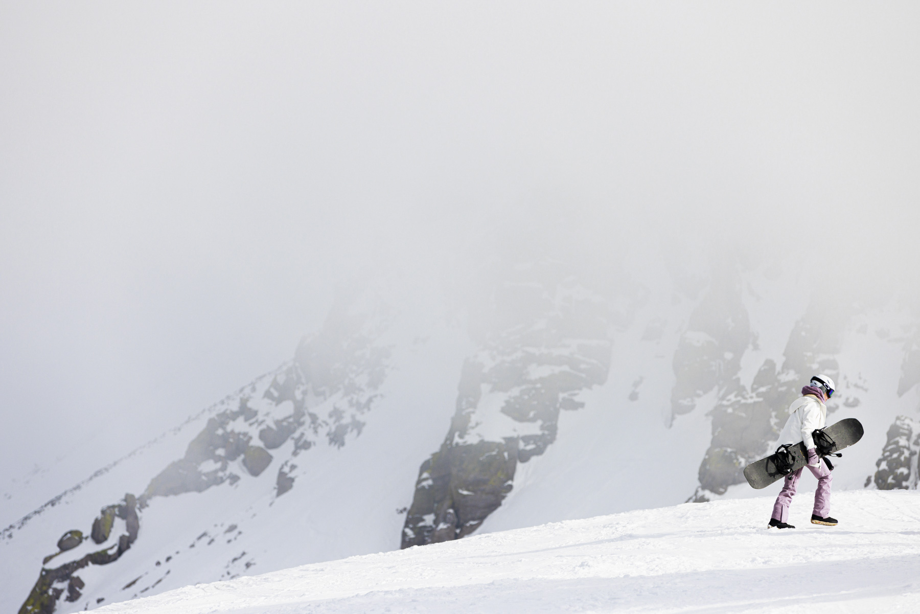 A snowboarder dressed in winter gear walks uphill on a snowy Idaho mountain slope, carrying a snowboard. Rocky, snow-covered peaks are partially visible through mist in the background, capturing the spirit of winter sports.