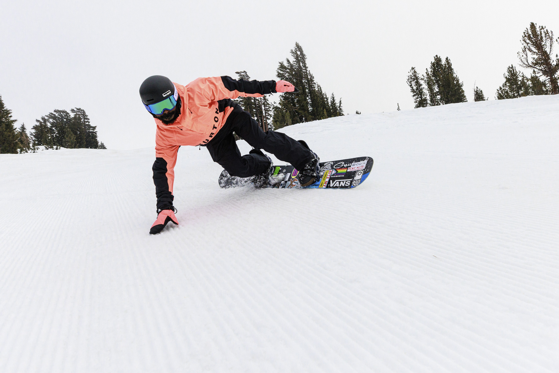 A snowboarder wearing a pink jacket and black helmet carves sharply on a snowy slope, showing off their winter sports skills as one hand touches the ground. Captured by a sports apparel photographer, trees are visible in the background.