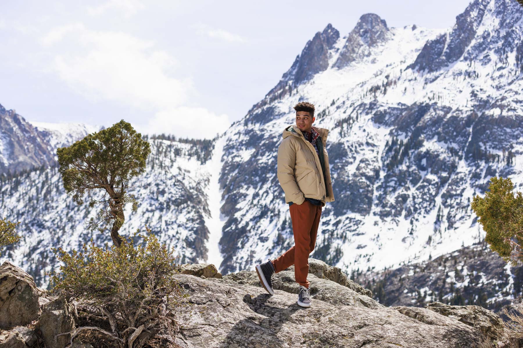 A person wearing a beige jacket, red pants, and gray shoes stands on a rocky ledge with snow-covered mountains behind them—an ideal shot for a sports apparel photographer or a Dicks Sporting Goods winter sports campaign.