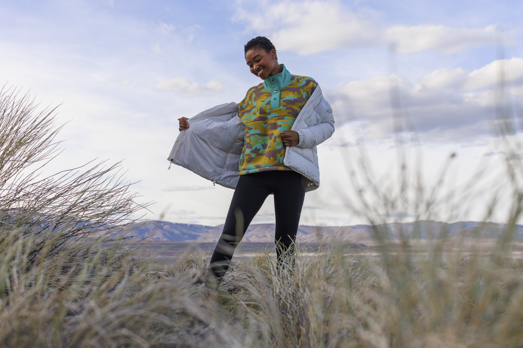 A person stands in tall grass outdoors in Idaho, smiling and holding open a light-colored jacket to reveal a colorful patterned top, with mountains and a cloudy sky in the background—captured by a skilled commercial photographer.