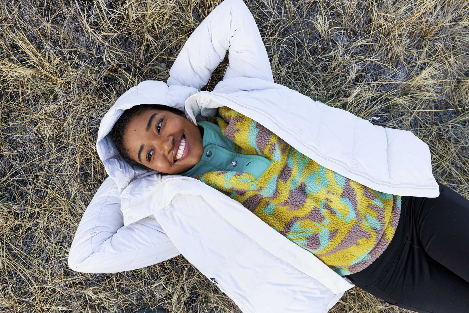 A person lies on dry grass in Sun Valley, Idaho, smiling up at the camera. They wear a white puffer jacket, a colorful fleece top, and black pants—perfect for a sports apparel photographer capturing relaxed outdoor vibes.
