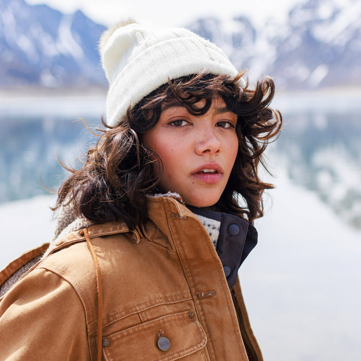 A person with curly hair, dressed in a white knit beanie and brown jacket, stands outdoors by a lake in Idaho, snowy mountains blurred behind—captured with the keen eye of a commercial photographer.