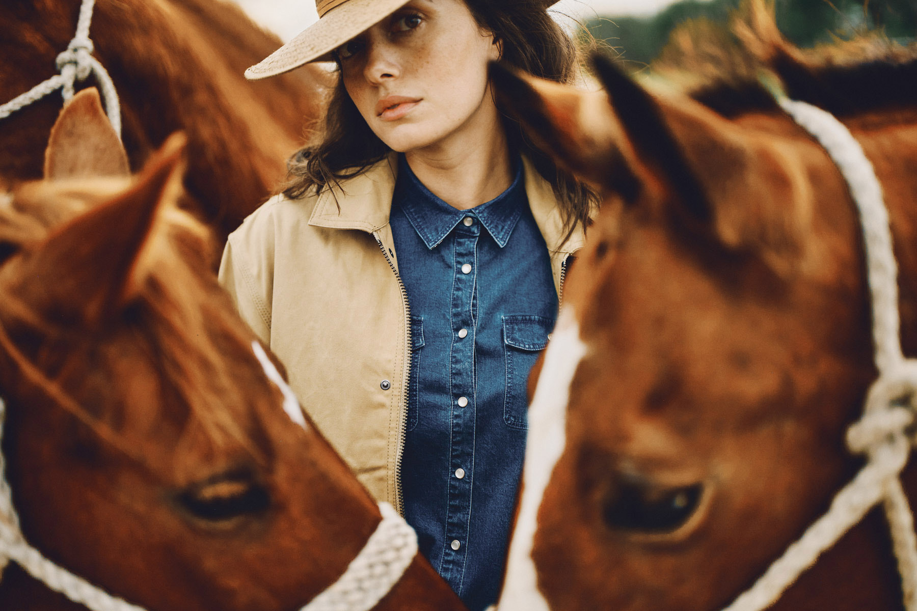 A woman wearing a hat and denim shirt stands among two brown horses with white rope halters, looking thoughtfully at the camera. The outdoor scene evokes King Ranch vibes, suggesting a classic country setting captured by a fashion photographer.