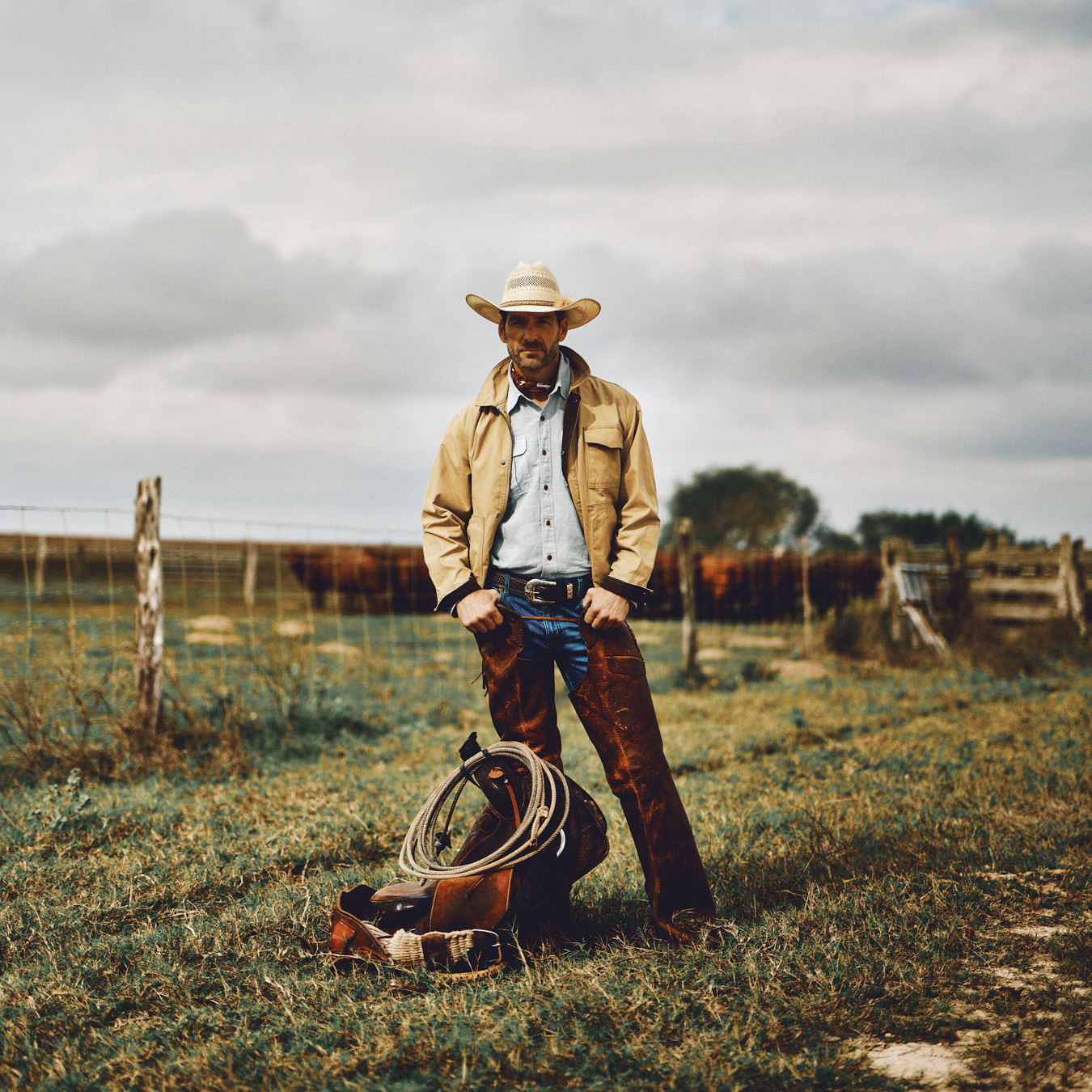 A cowboy wearing a hat and boots stands in a grassy Texas field beside a fence, with coiled rope and leather gear at his feet. Cows and cloudy skies fill the background, capturing a classic Western scene.
