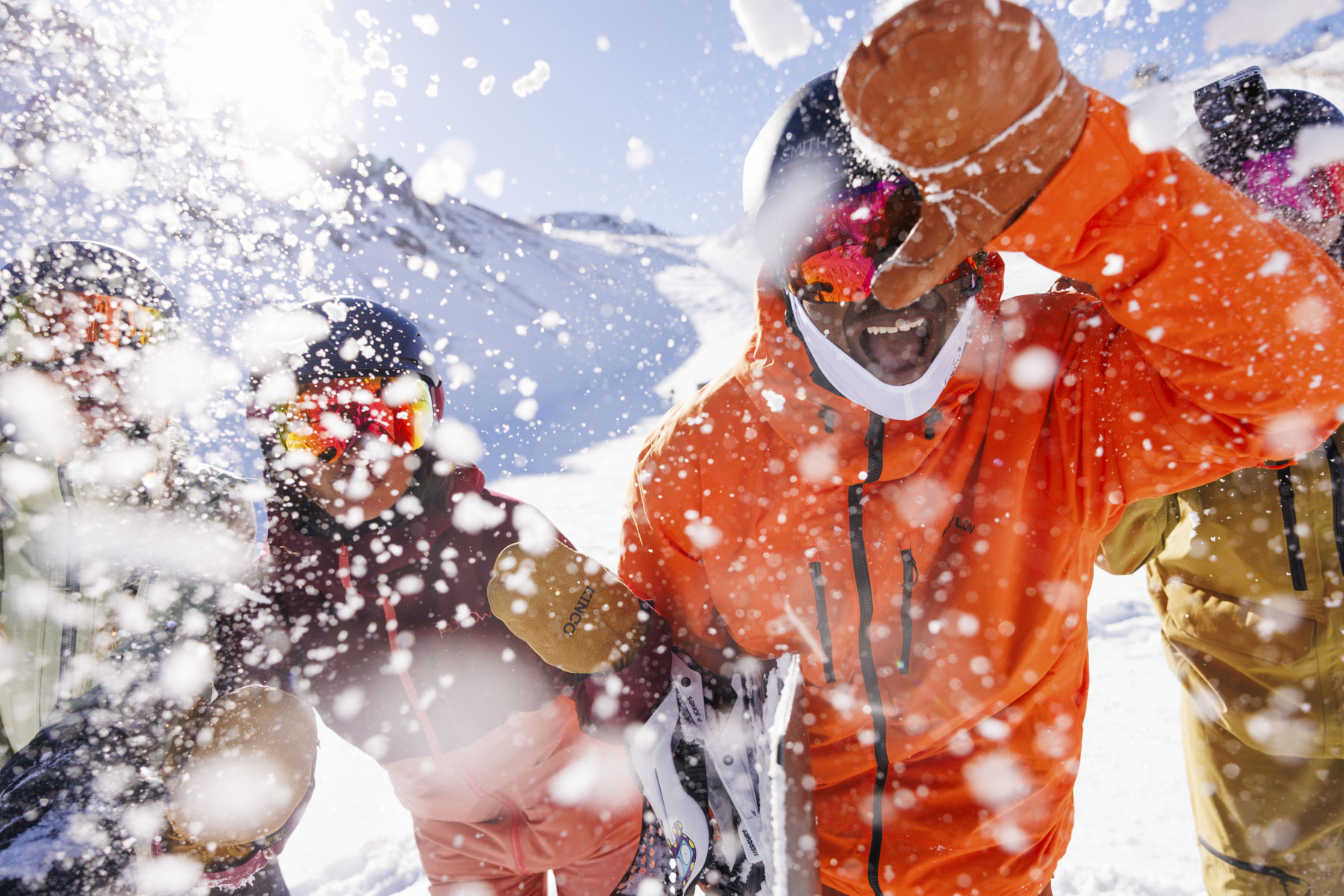 Four people in colorful winter apparel laugh as snow flies into the camera on a sunny mountain slope, enjoying a lively moment of adventure while skiing or snowboarding.