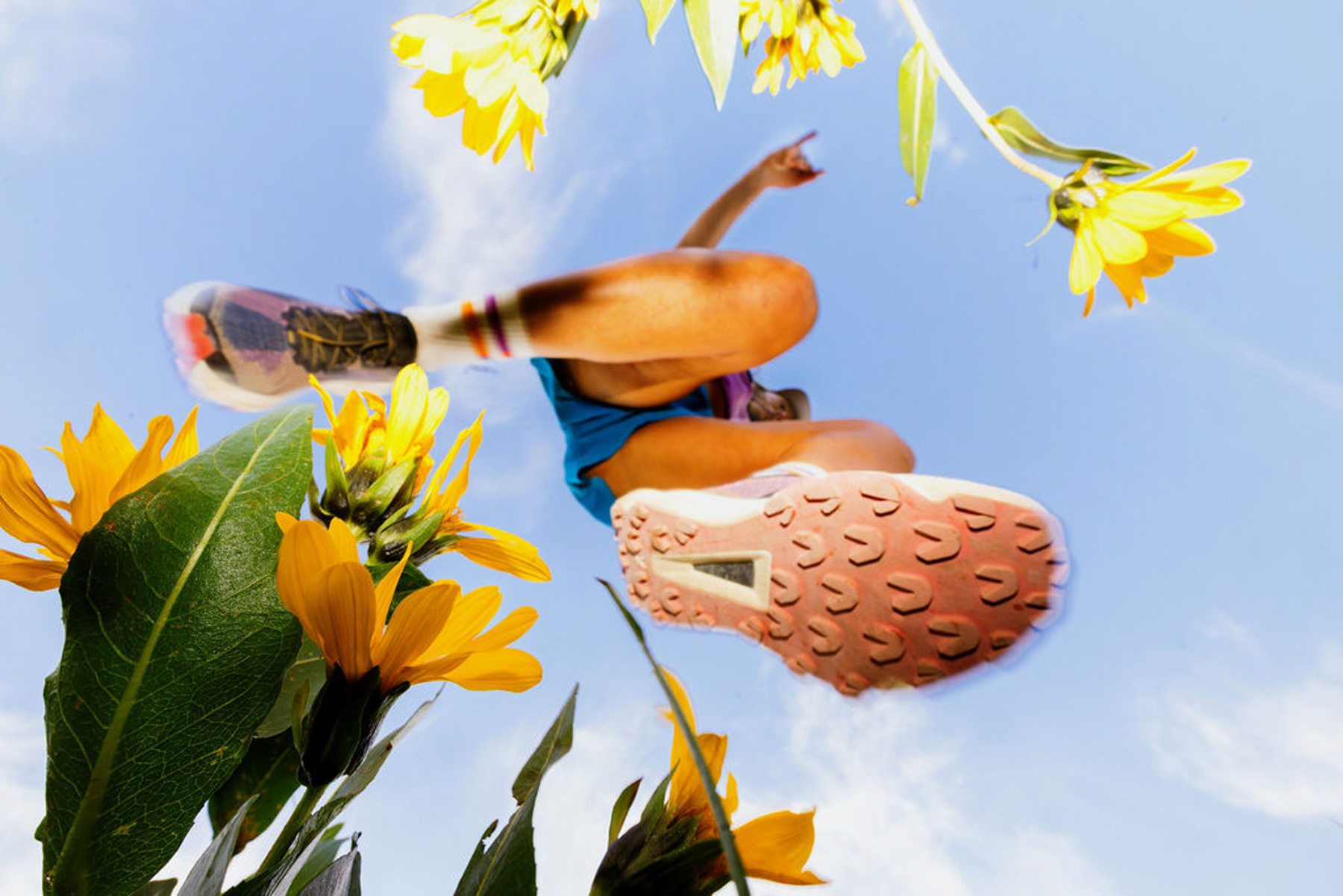 Low-angle view of a person running outdoors above yellow sunflowers, with blue sky in the background. The runner’s shoes and legs are prominent, creating a dynamic, energetic scene. Photo by Sofia Jaramillo.