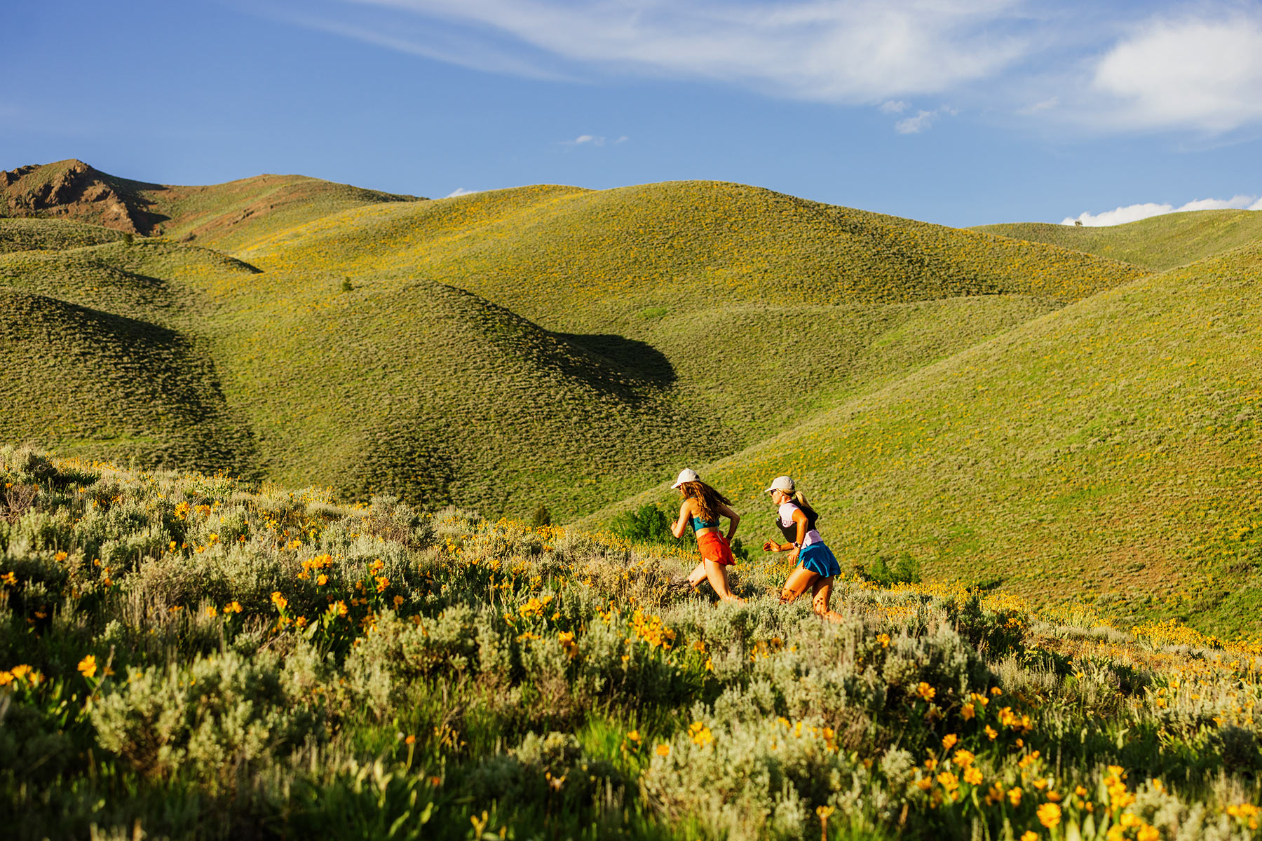 Two people wearing hats and athletic clothing run up a wildflower-covered hillside, embracing adventure outdoors, with rolling green hills and a blue sky in the background.
