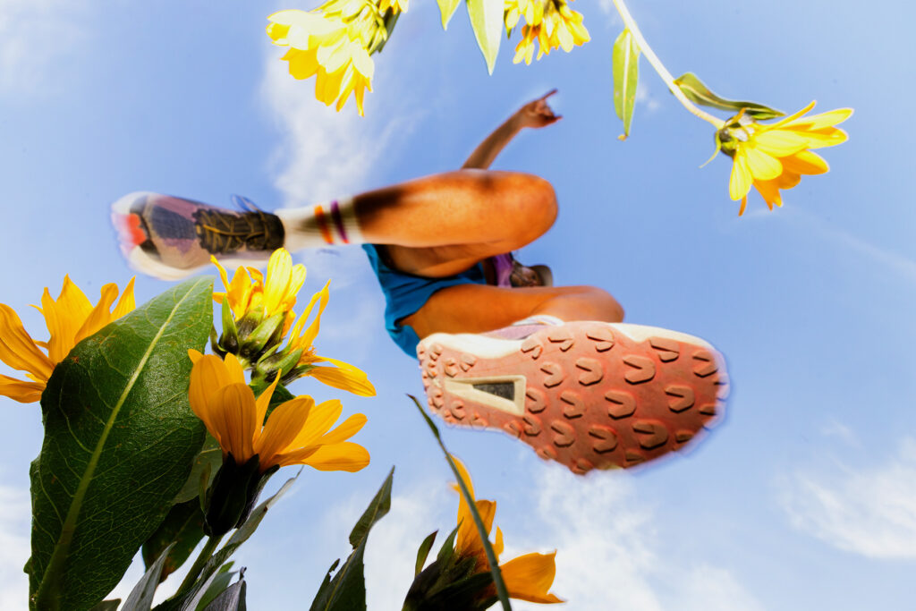 Low-angle view of a runner mid-jump above yellow flowers, with the sky and clouds in the background. The focus on the runner’s shoe and legs highlights action, movement, and the adventure of trail running outdoors.