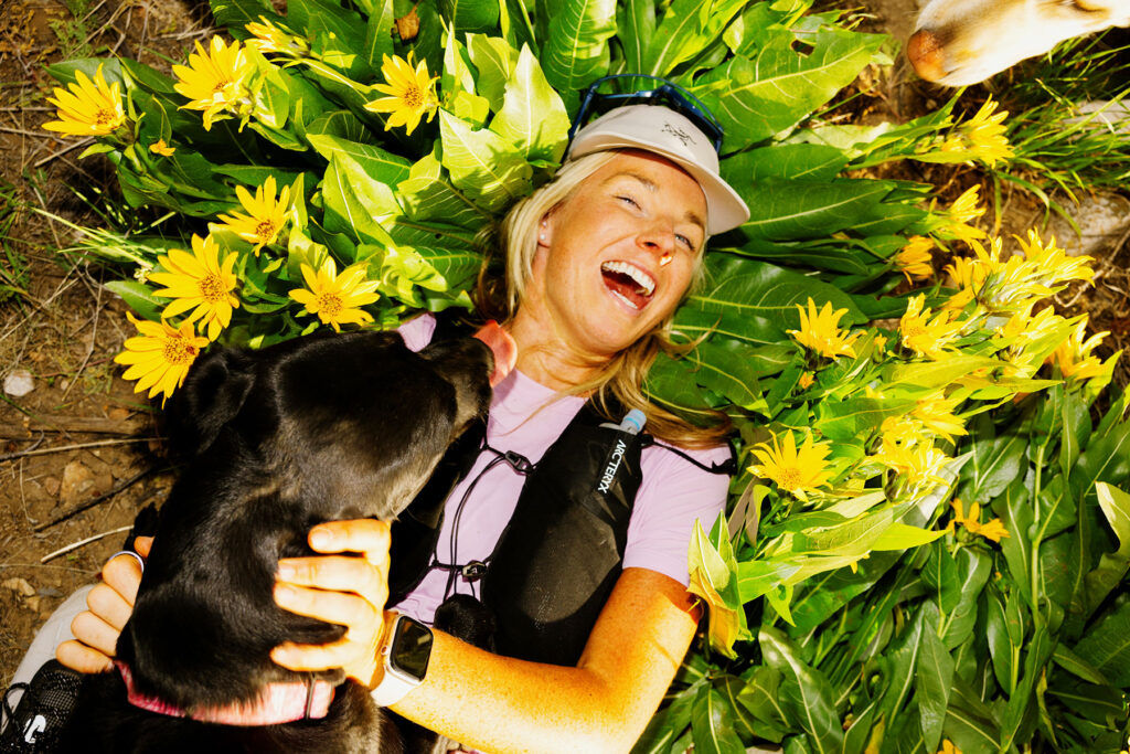 A woman lies in a patch of yellow flowers, laughing as a black dog licks her face. Shot by commercial photographer Sofia Jaramillo, she wears a hat, sunglasses, and a smartwatch, appearing joyful and relaxed in the sunny outdoor setting.