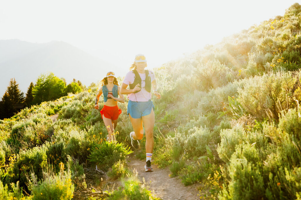Two people enjoy trail running on a sunlit dirt path winding through green, grassy hills and mountains. Wearing athletic gear and hats, they embrace an outdoor adventure in bright natural light—perfect for a commercial photographer to capture.