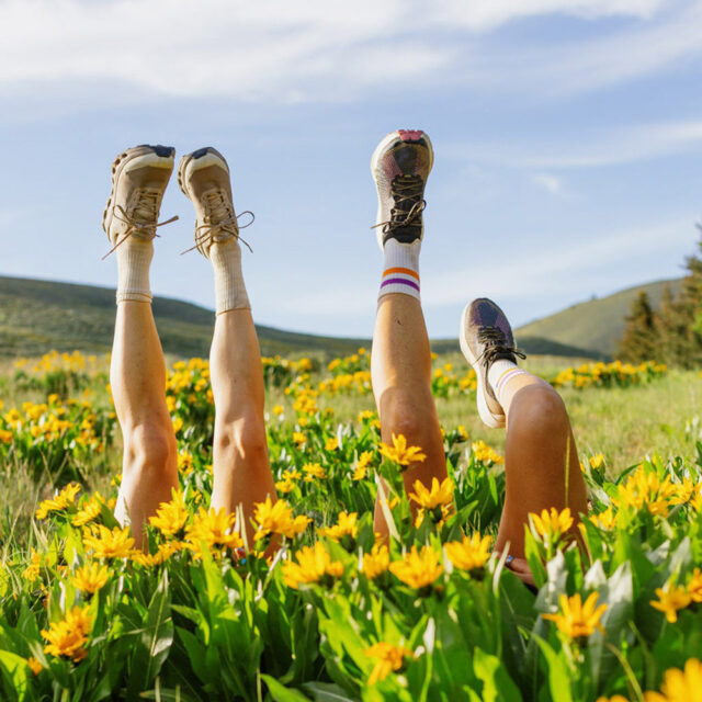 Three people lying on their backs outdoors in a field of yellow wildflowers, legs playfully raised in the air, wearing sneakers and colorful socks under a blue sky with hills beyond. Photo by Sofia Jaramillo.