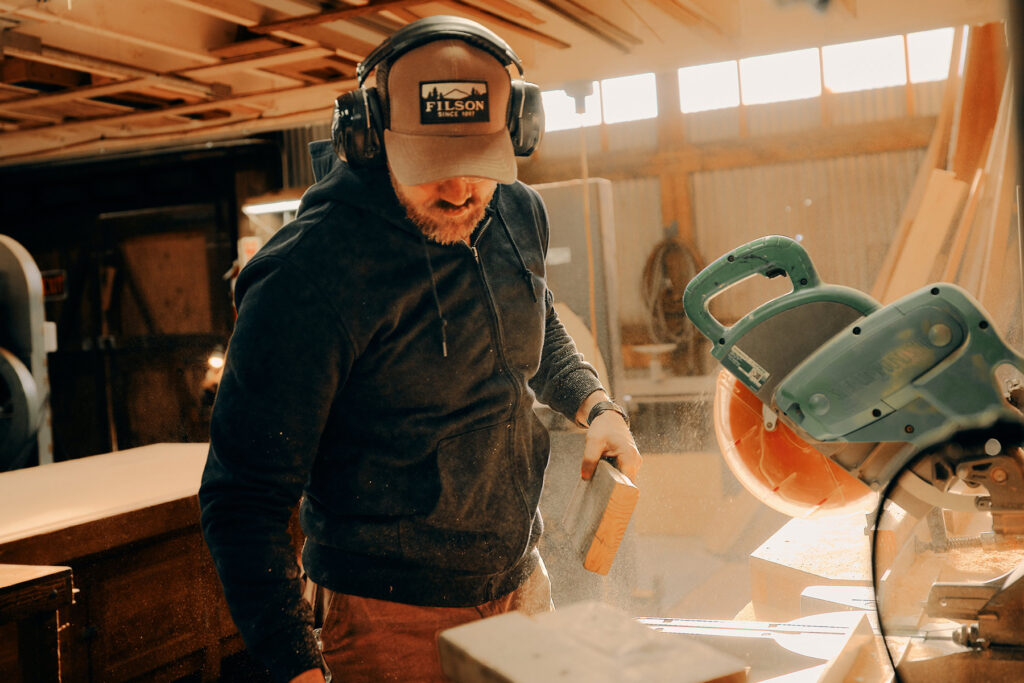 A man wearing a Filson cap, safety glasses, and ear protection is working in a sunlit woodshop, sanding a piece of wood near a circular saw, with sawdust in the air.
