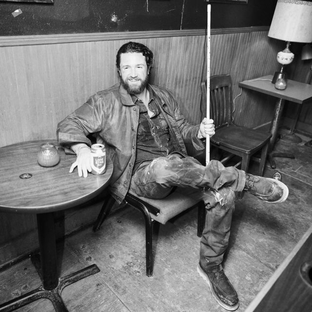 A man with a beard sits casually on a chair in a bar, holding a pool cue and a can of beer. Showing off rugged Tecovas style, he wears a leather jacket and worn jeans—like he’s ready for his next fashion photographer shoot.