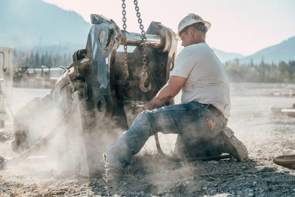 A construction worker in rugged workwear and a hard hat kneels on dusty ground, operating heavy machinery outdoors with mountains in the background. Chains hang from the equipment as sunlight filters through the dust, reminiscent of Travis Gilletts dynamic photographic style.