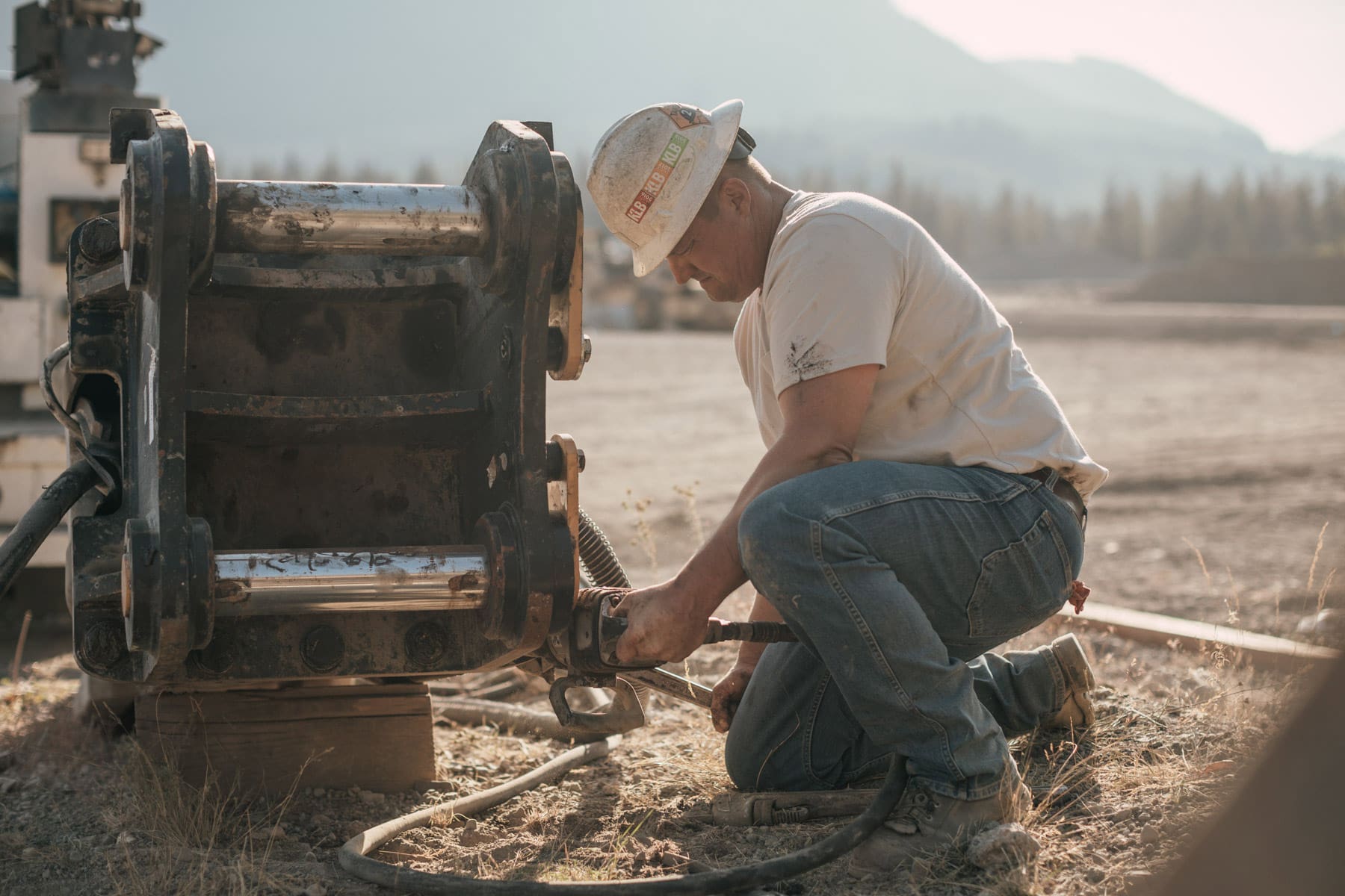 A construction worker wearing a hard hat and Levis jeans kneels on the ground, using a tool to fix a large piece of industrial machinery outdoors, with mountains and trees visible in the background.