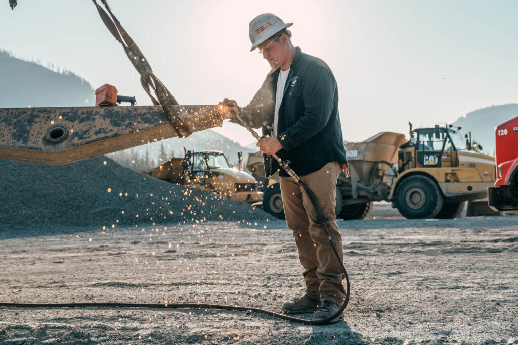 A construction worker in workwear uses a tool that creates bright sparks, standing outdoors on a gravel lot with heavy machinery and mountains in the background, captured by a commercial photographer.