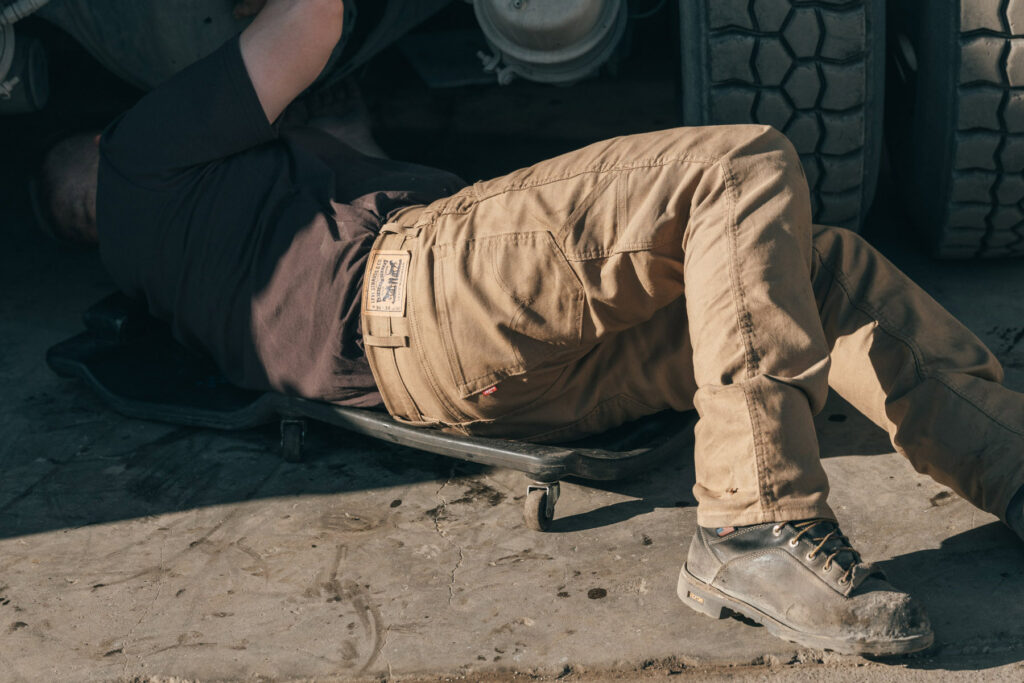 A person in work boots and tan pants lies on a mechanic creeper under a large vehicle, performing maintenance or repair work in a Seattle garage—captured with the keen eye of fashion photographer Travis Gillett.