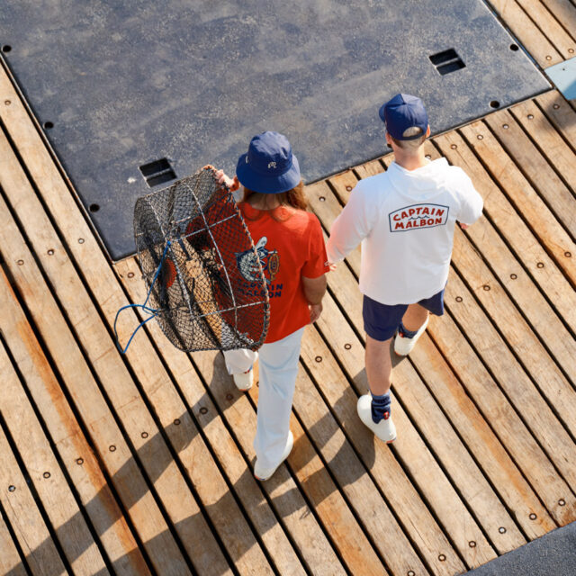 Two people walk on a wooden boat dock; one in a red shirt and blue hat carries a crab trap, while the other wears a white shirt reading CAPTAIN MALBON, blue shorts, and a blue cap.