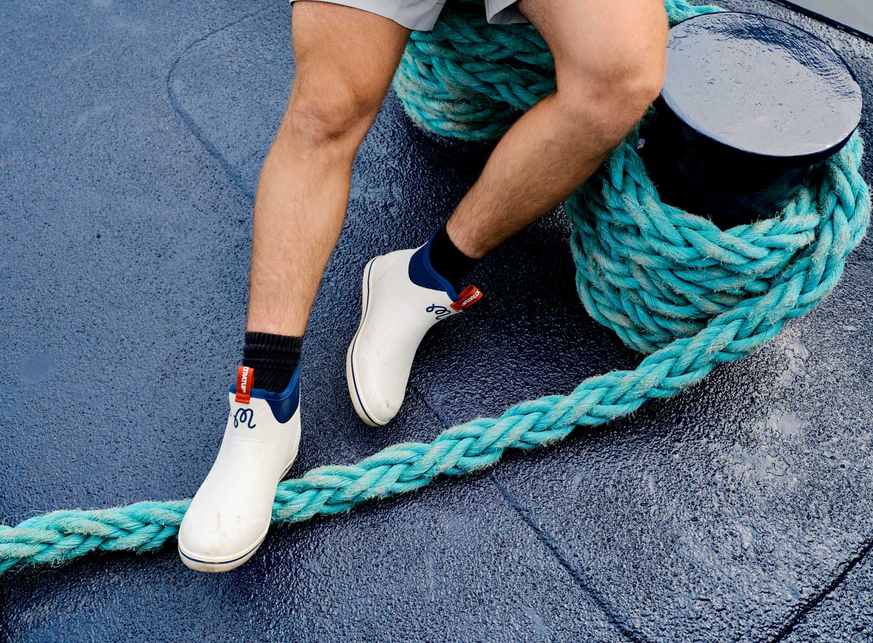A person, captured by fashion photographer Travis Gillett, wears white slip-on boots and black socks from Xtratuf and Malbon Golf, standing on a wet boat deck next to a thick coiled turquoise rope and black mooring bollard. Only the lower legs and feet are visible—a nod to practical workwear.