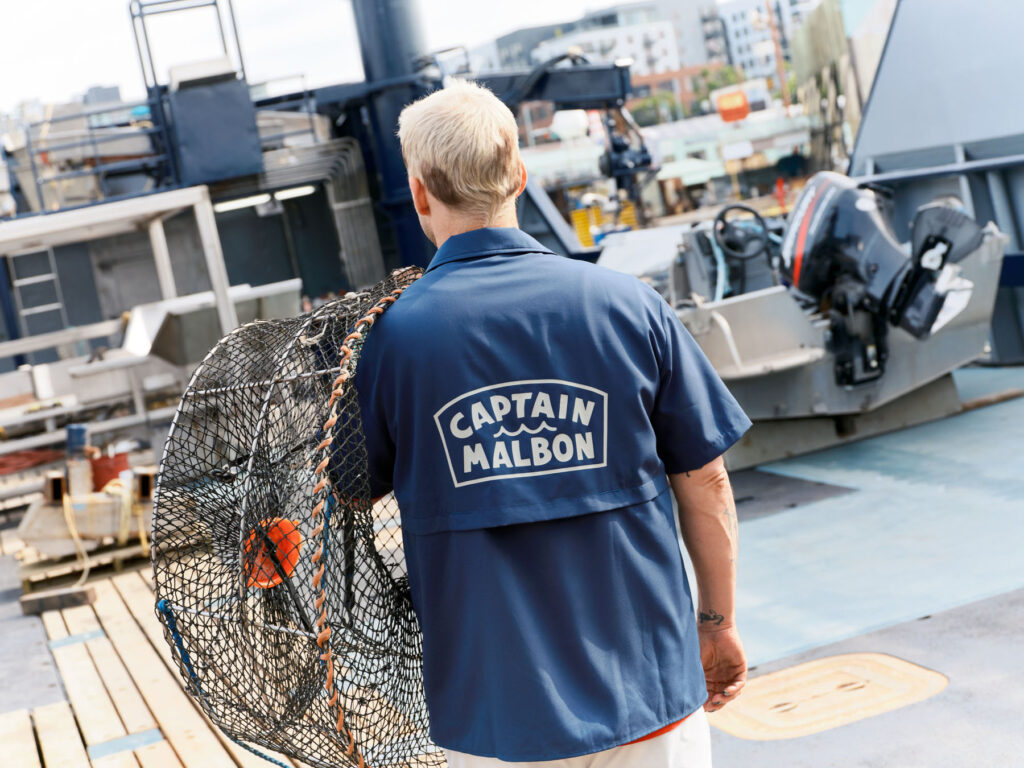 A person with short blond hair, wearing a blue workwear shirt labeled Captain Malbon, carries a large fishing net on their shoulder while standing on a docked boat. Buildings are visible in the background. Shot by a Seattle commercial photographer.