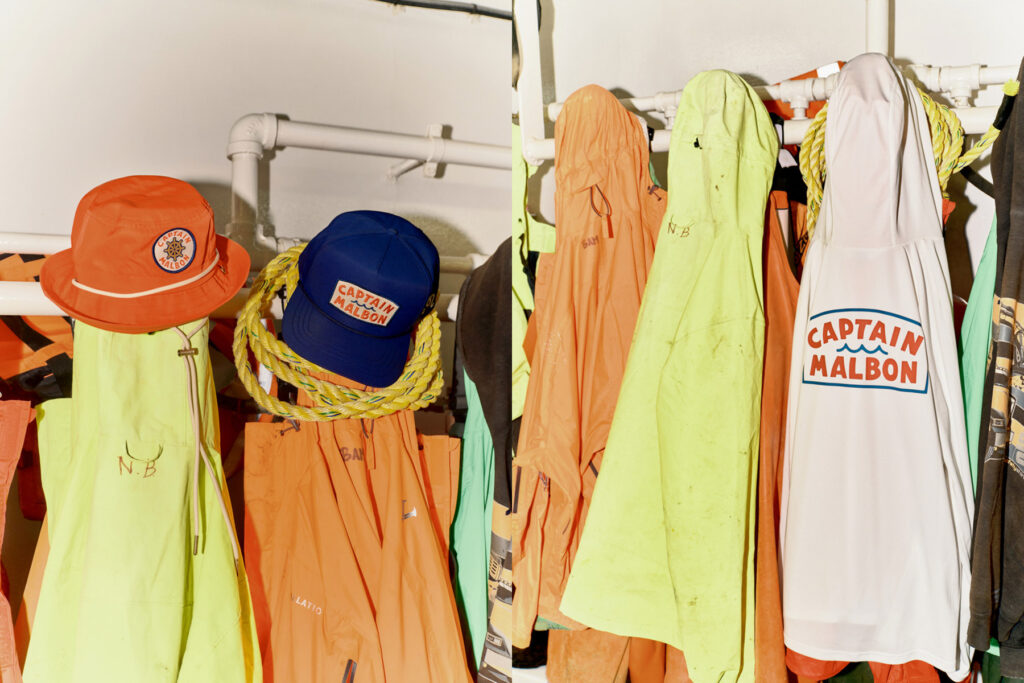 Brightly colored jackets, hats, and a hoodie hang on hooks. An orange bucket hat and blue cap say “Captain Malbon,” while a white hoodie reads “Captain Malbon.” Captured by a Seattle commercial photographer for Malbon Golf. Yellow rope hangs above the clothing.