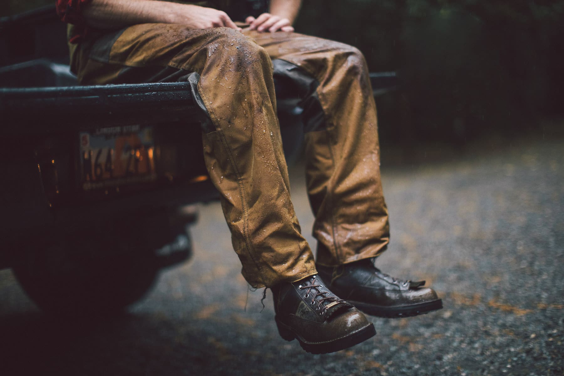 A person wearing brown pants and black boots sits on the edge of a vehicle’s tailgate in the rain, water droplets visible—a striking moment captured by fashion photographer Travis Gillett.