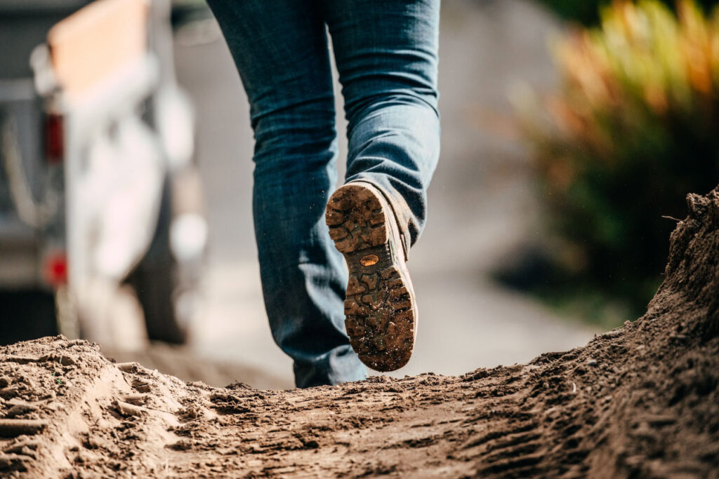 A person in blue jeans and sturdy workwear walks on a dirt path, seen from behind at ground level, with focus on the sole of their shoe and textured dirt beneath their feet. Blurred greenery is visible in the background.