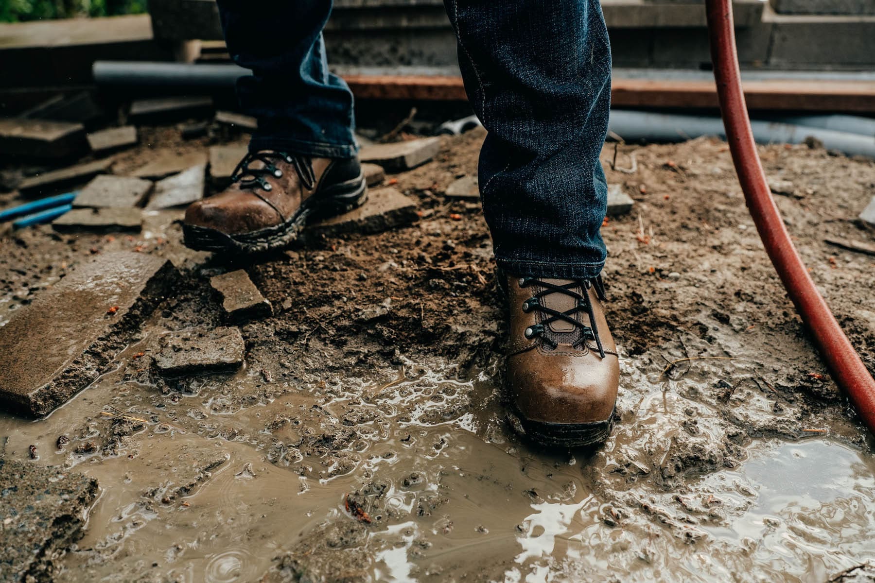 A person wearing brown work boots and jeans stands on muddy, wet ground surrounded by broken pieces of concrete and a red hose—captured by a Seattle commercial photographer.