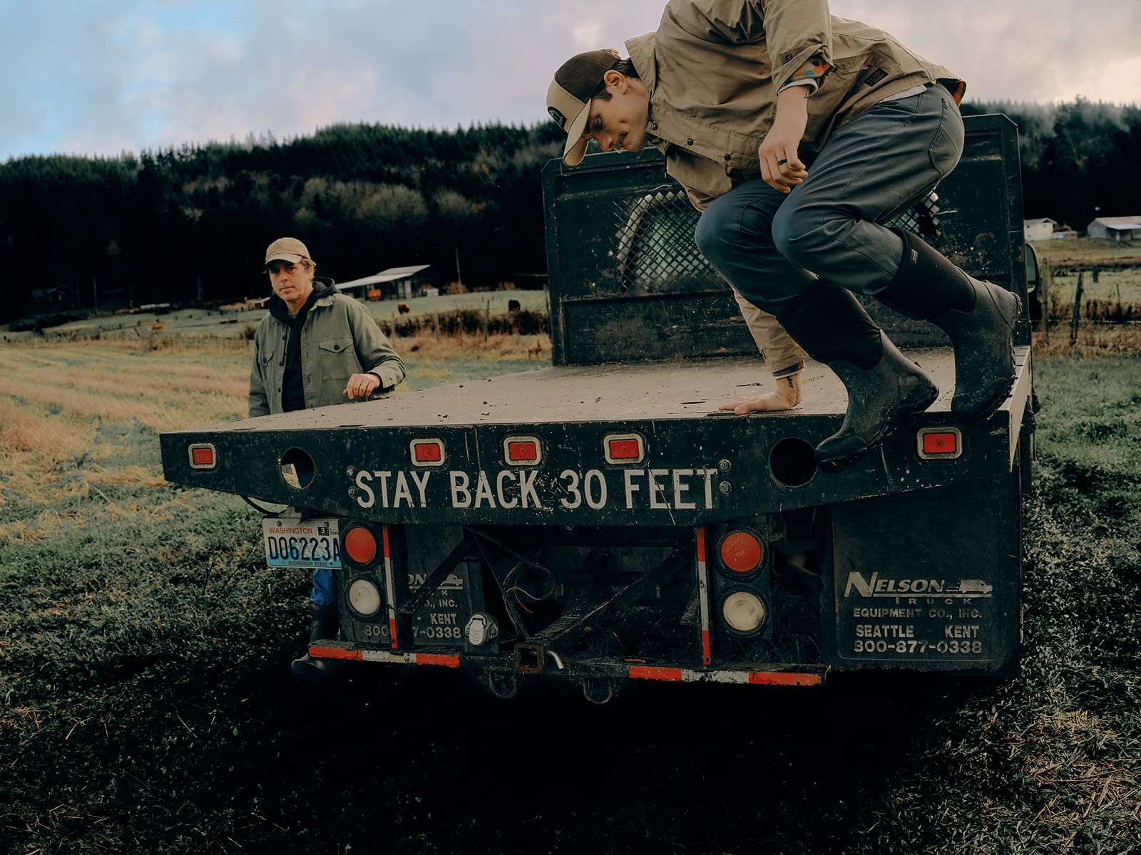 Two people in industrial work clothes stand on the back of a flatbed truck in a grassy field near Seattle; one steps off while the other watches. The truck bed reads STAY BACK 30 FEET. Trees and a building are visible in the background.