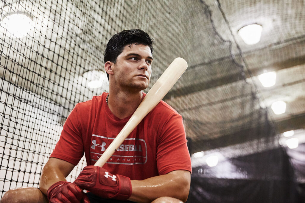 Baseball player Andrew Benintendi wearing a red Under Armour shirt and red Under Armour gloves sits on a bench indoors, holding a bat on his shoulder, with netting and bright lights in the background.