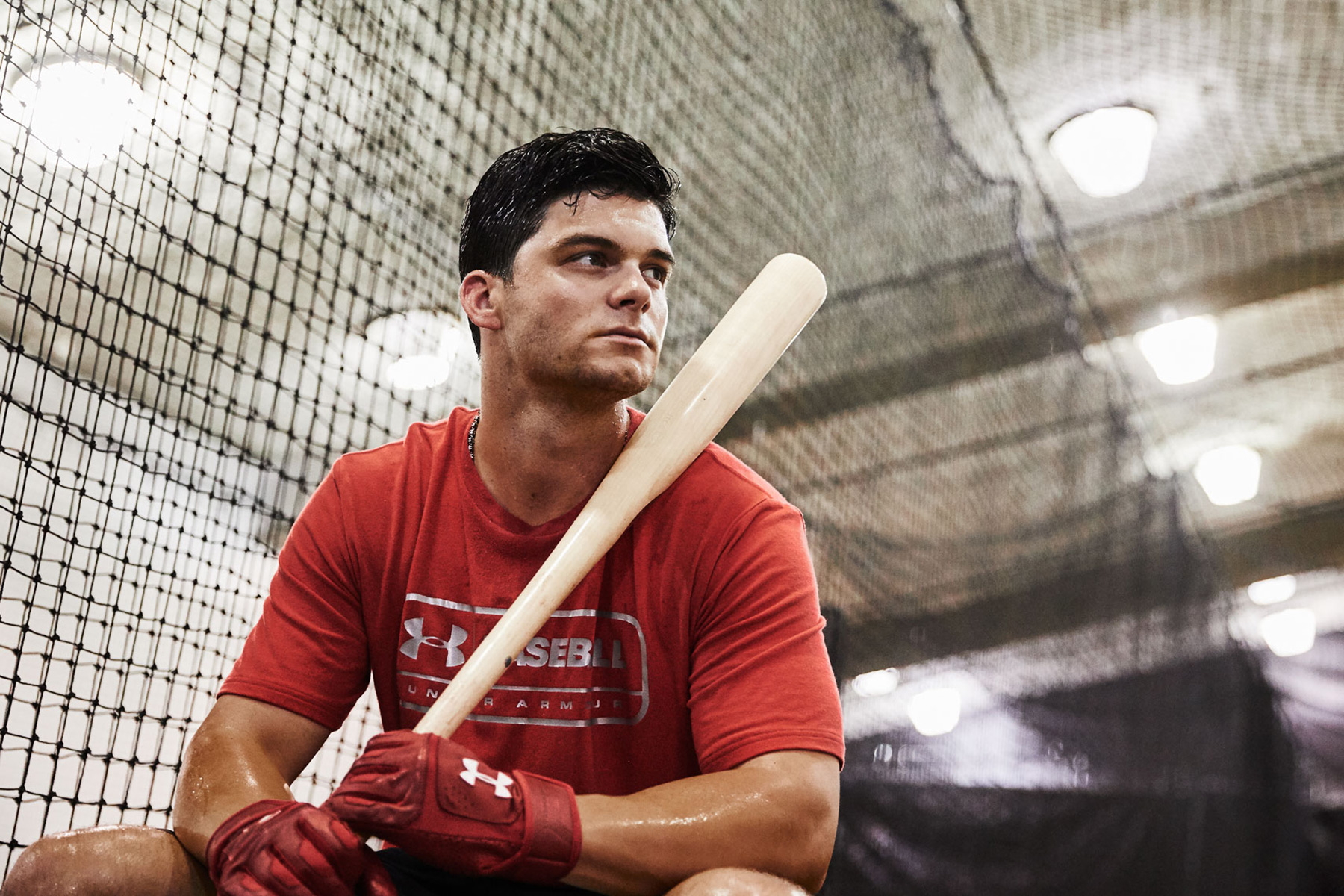 Baseball player Andrew Benintendi wearing a red Under Armour shirt and red Under Armour gloves sits on a bench indoors, holding a bat on his shoulder, with netting and bright lights in the background.