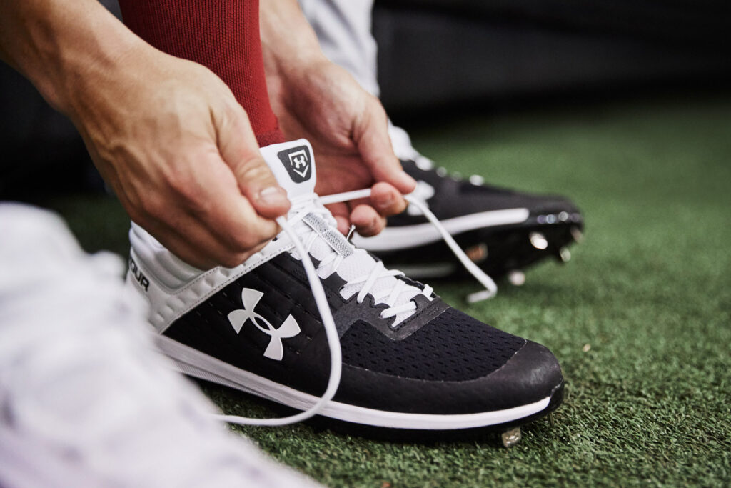 Close-up of a person tying the laces on black and white Under Armour athletic cleats while standing on artificial turf.