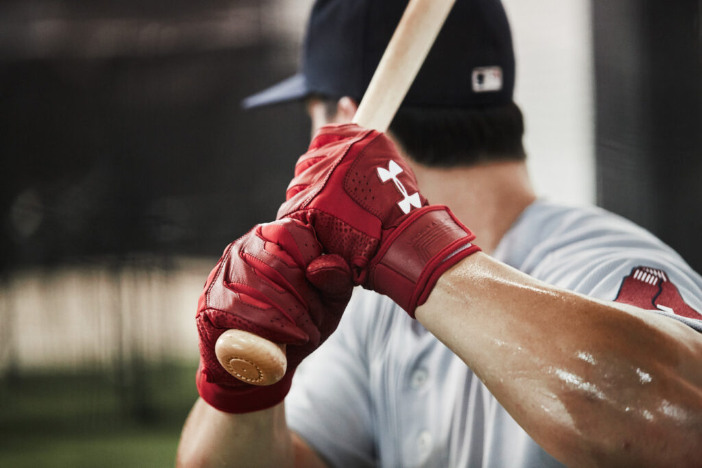 Baseball player Andrew Benintendi wearing a red Under Armour shirt and red Under Armour gloves sits on a bench indoors, holding a bat on his shoulder, with netting and bright lights in the background. Fj Hughes, a Baltimore based advertising and sports photographer worked with him on a campaign for Under Armour. 