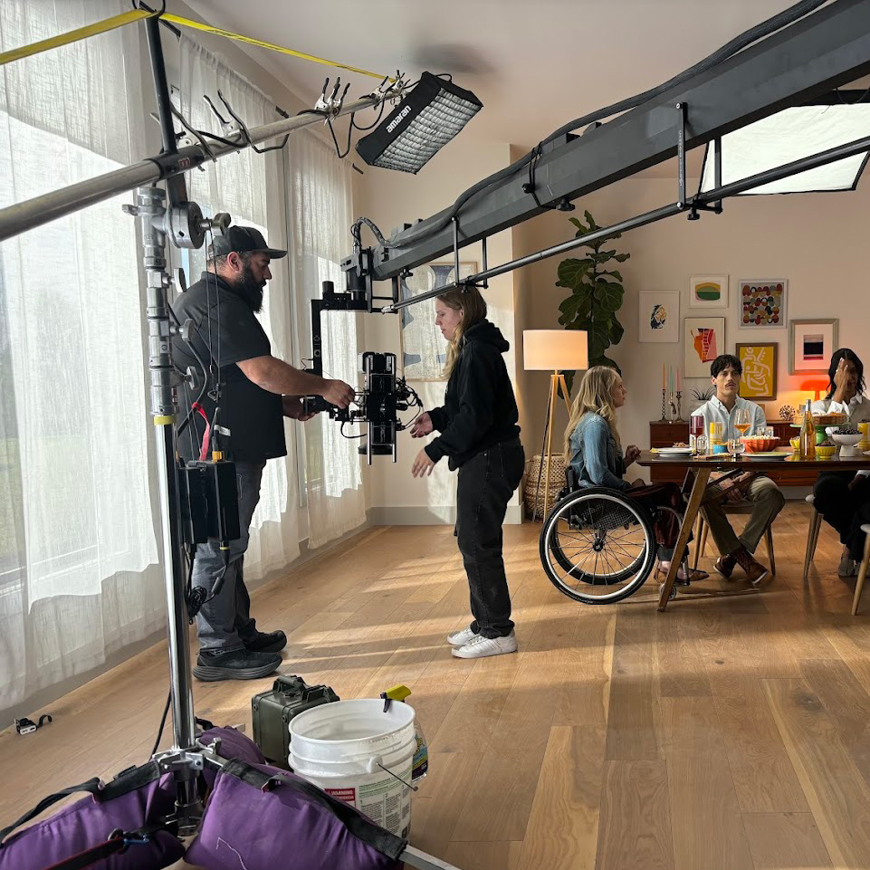 A film crew sets up a camera on a large rig in a bright living room, capturing footage of three people, including one in a wheelchair, at a dining table—perfect for showcasing expert post production services.