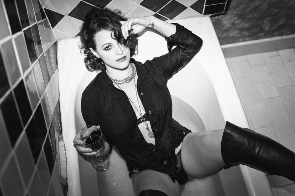 Actress Clementine Heath wears a black shirt and thigh-high boots as she sits in a bathtub, holding a glass of red wine. Her hair is styled in loose curls, and she wears layered necklaces. The image is in black and white.