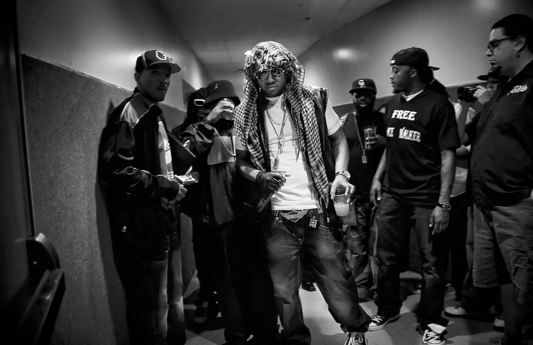 A group of Baltimore rappers sit in a a dimly lit hallway before going on stage. Rapper Bossman, in front, wears sunglasses, a headscarf, and holds a drink, while others gather around him, some looking towards the camera.