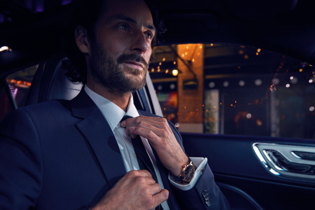 A man in a suit sits in a car at night, adjusting his tie and looking confident. Warm city lights are visible outside the window, creating a sophisticated and stylish atmosphere.