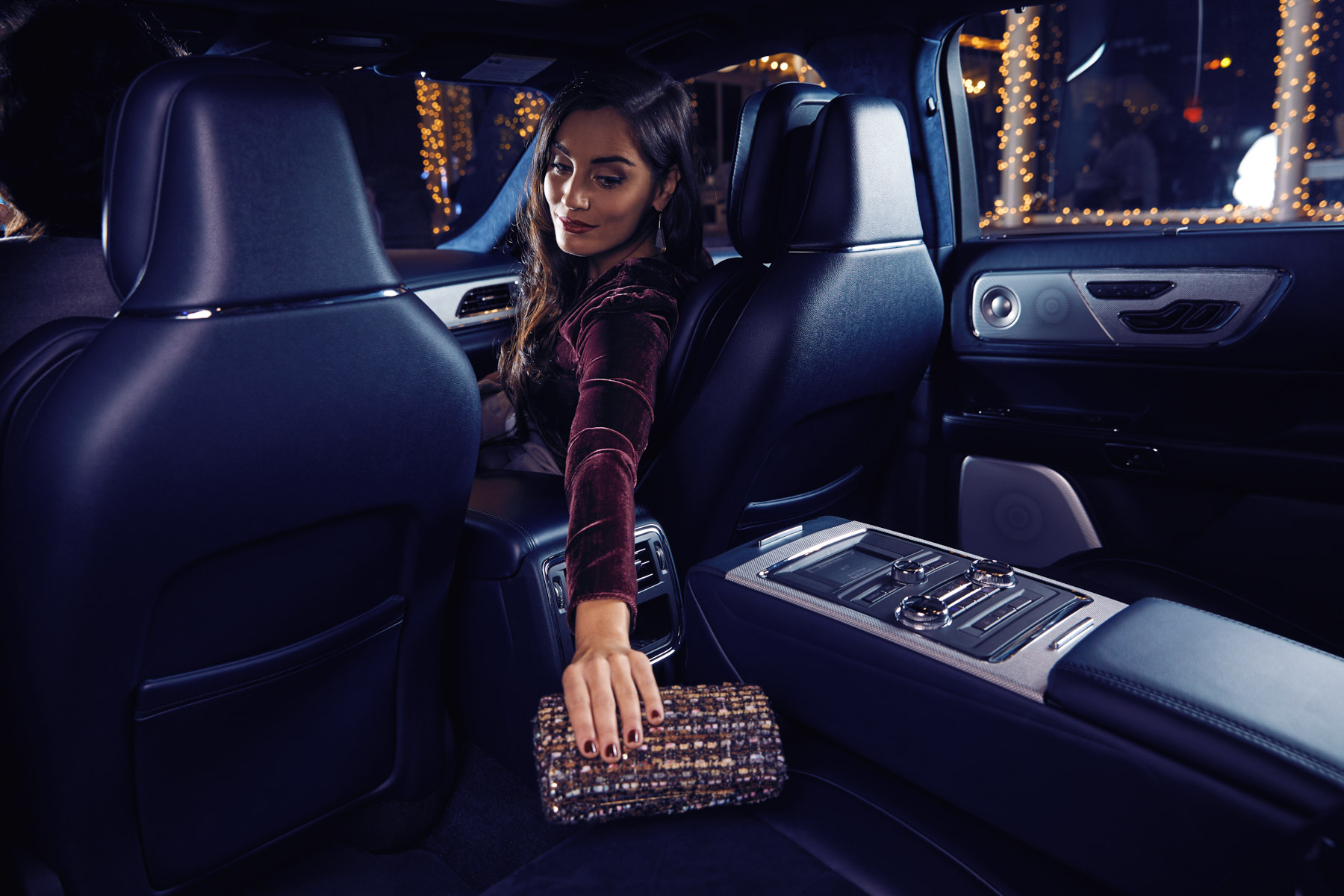 A woman sitting in the back seat of a luxury car at night, reaching for a sparkling clutch on the center armrest; city lights and reflections are visible outside the windows.
