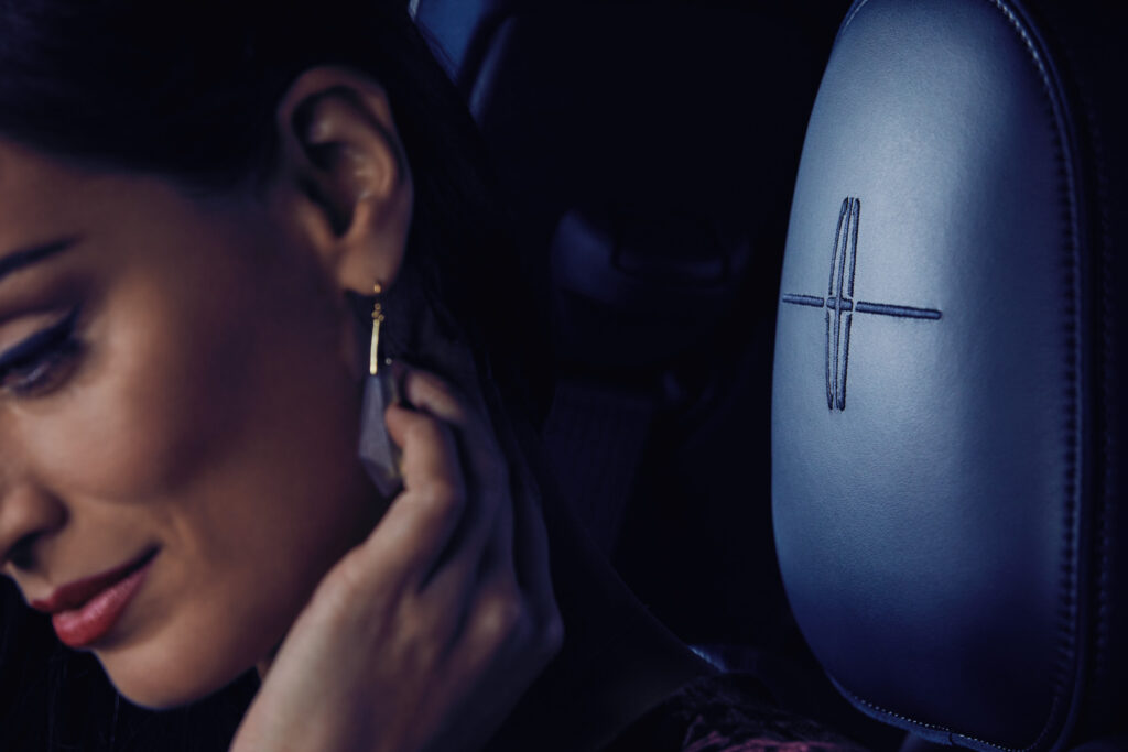 A woman touches her neck while sitting in a car, with the leather headrest embroidered with the Lincoln logo prominently visible beside her.