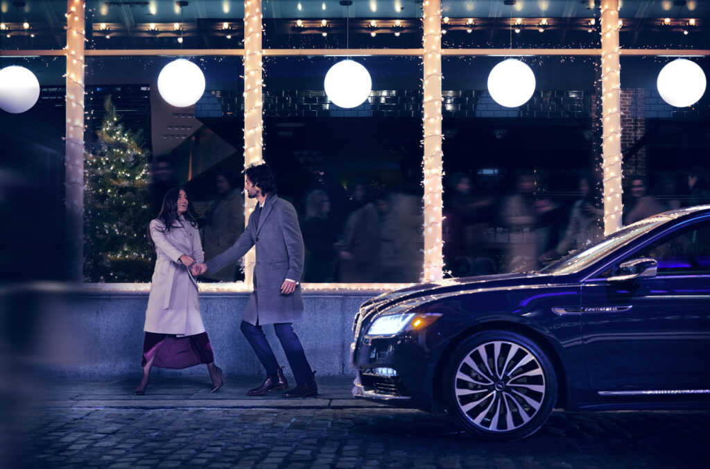 A couple in winter coats hold hands and walk on a city sidewalk at night, passing a decorated Christmas tree and festive lights, with a parked luxury car in the foreground.