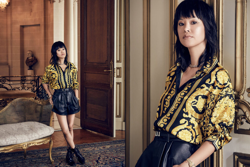 A woman with straight dark hair wears a gold and black patterned shirt tucked into high-waisted black leather shorts, standing confidently indoors by ornate doors and furniture.