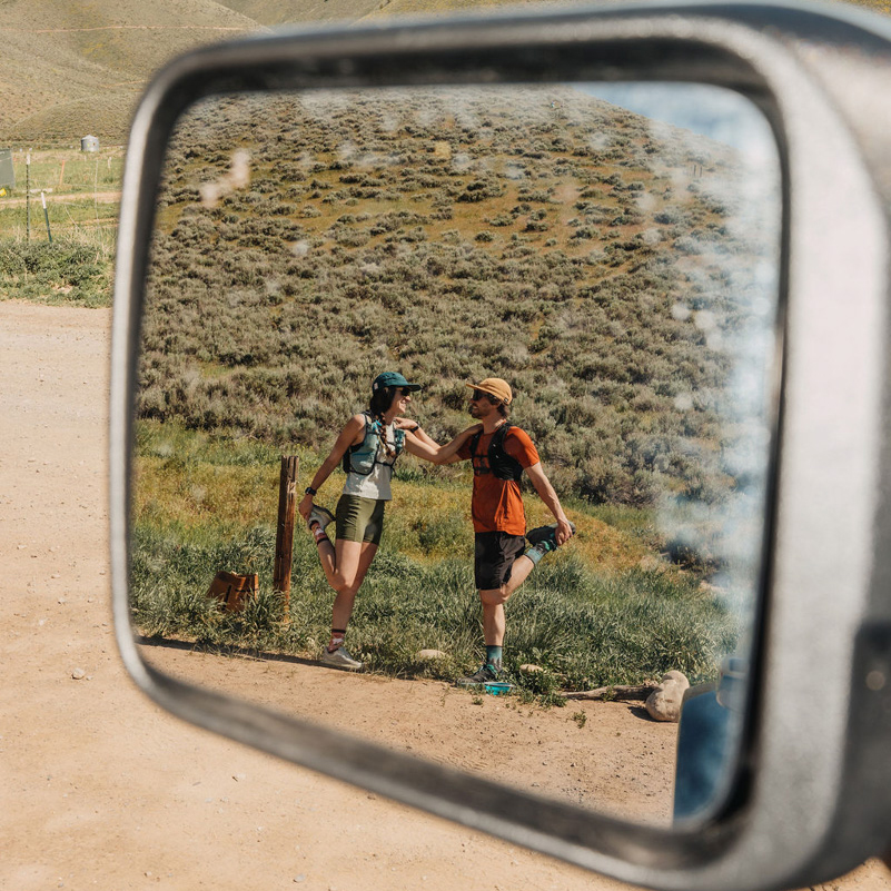 A reflection in a car side mirror shows two trail runners in athletic gear stretching and smiling at each other on a dirt trail with grassy hills in the background.