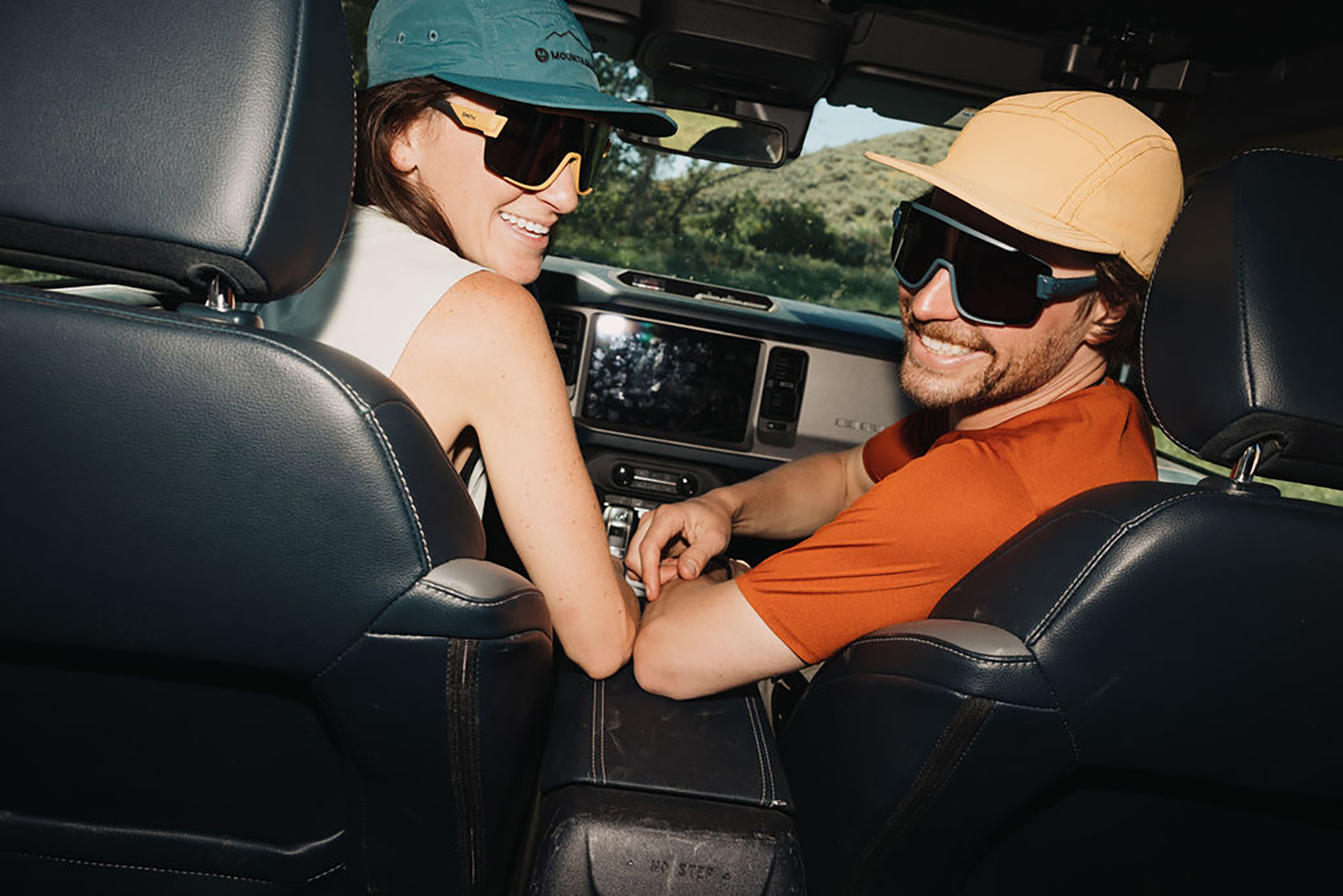 A smiling man and woman wearing sunglasses and hats sit in the front seats of a car, looking back toward the camera. Sunlight shines through the windows, and greenery is visible outside.