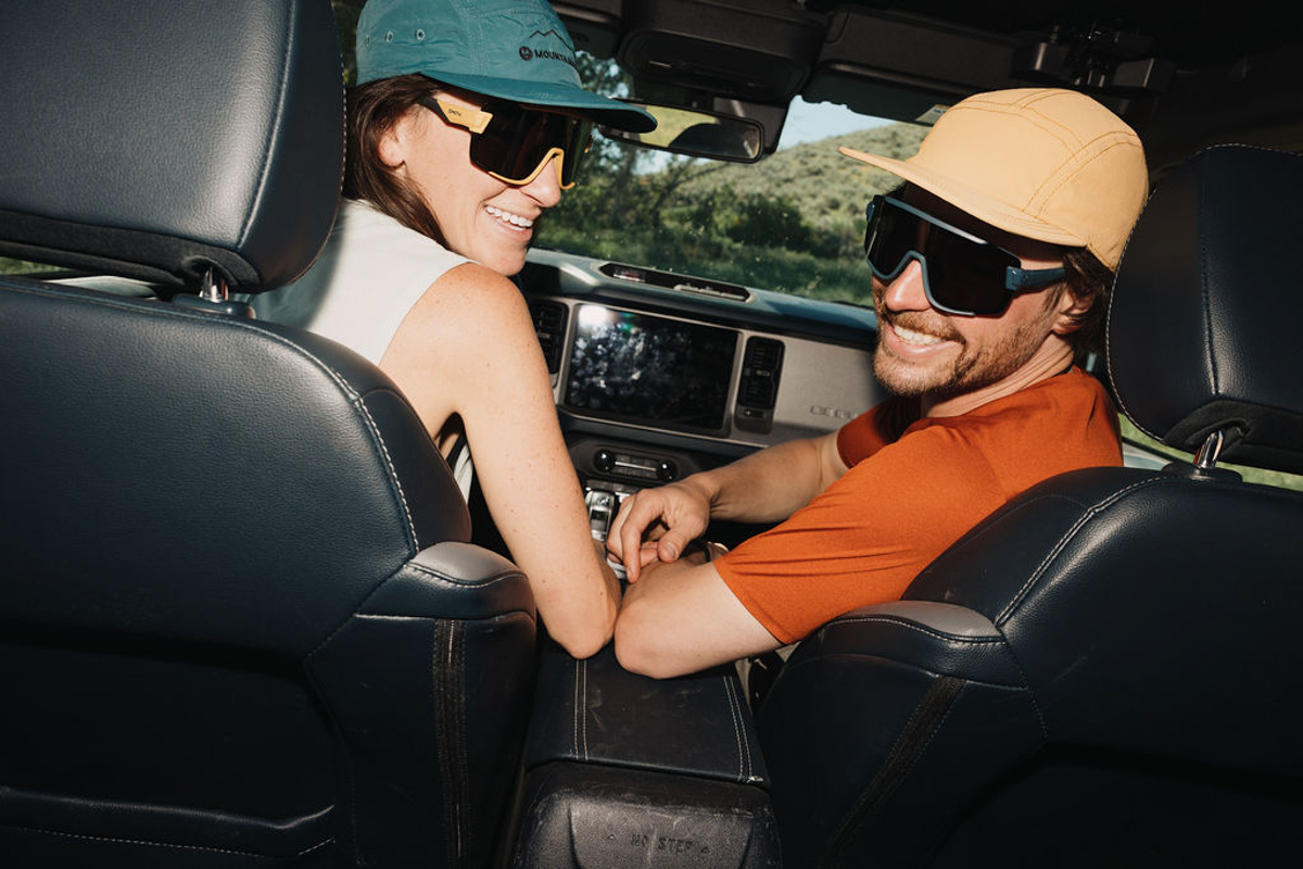 Two people wearing sunglasses and hats sit in the front seats of a car, smiling and looking back toward the camera. Sunlight streams in, and green scenery is visible through the windows right as they arrive for a trail run.