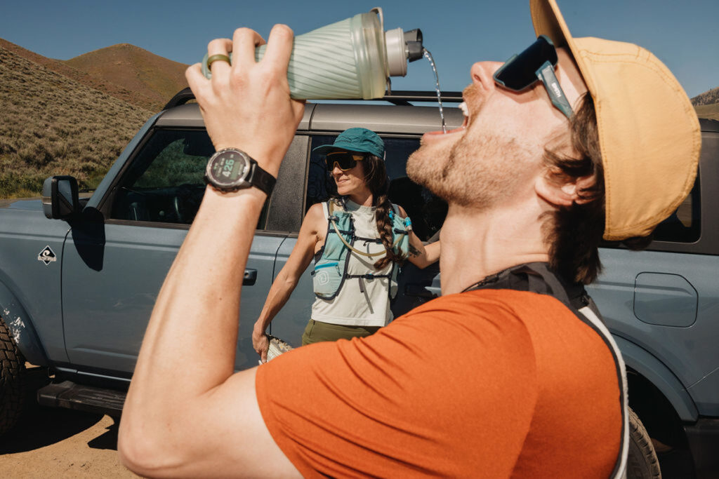 A man in sunglasses and an orange shirt drinks from a water bottle near a Ford Bronco SUV, while a woman in a blue hat and sunglasses stands by the vehicle, smiling and stretching before her tail run. They are in the great outdoors on a sunny day.